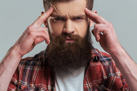 Handsome young bearded man in casual clothes is looking a camera and keeping fingers on forehead, on a gray background, close-upの写真素材