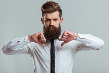 Handsome young bearded businessman in classic white shirt is showing not Ok sign, on a gray...