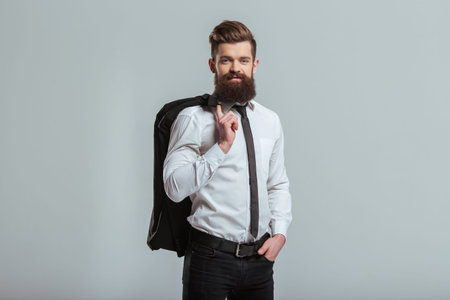 Handsome young bearded businessman in classic white shirt is holding a jacket on finger and smiling,...