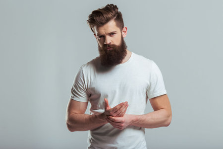 Handsome young bearded man is massaging his hand and looking at camera while standing against gray...