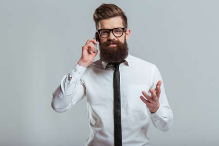 Handsome young bearded businessman in classic white shirt and eyeglasses is talking on the mobile phone, on a gray backgroundの写真素材