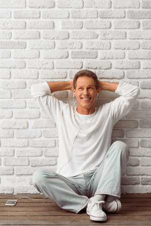 Handsome middle aged man in white sweatshirt is listening to music using a smart phone and smiling, sitting against white brick wallの写真素材
