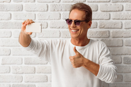 Handsome man in white sweatshirt and sunglasses is making a selfie, showing Ok sign and smiling,...