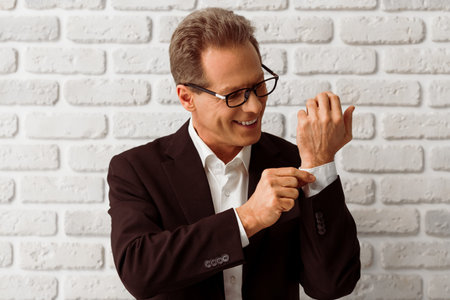 Handsome middle aged businessman in classic suit and eyeglasses is adjusting a cufflink and smiling,...