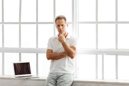 Thoughtful middle aged man in casual clothes is sitting near the window at home, laptop in the backgroundの写真素材