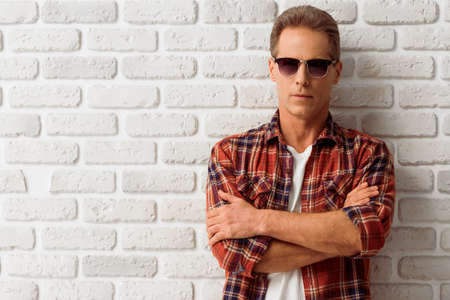 Handsome middle aged man in casual clothes and sunglasses is looking at camera while standing against white brick wallの写真素材