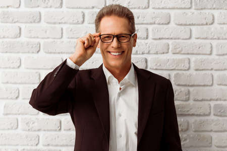 Handsome middle aged businessman in classic suit and eyeglasses is looking at camera and smiling, standing against white brick wallの写真素材