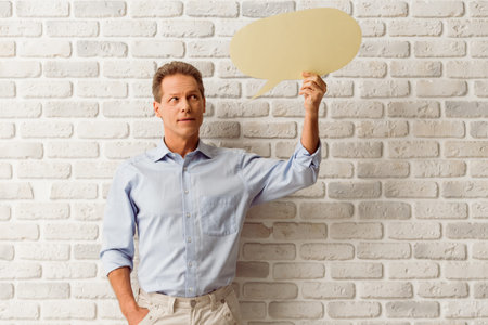 Handsome middle aged man is holding a yellow speech bubble and looking at it while standing against...