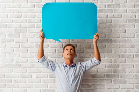 Handsome middle aged man is holding a blue speech bubble and looking at it while standing against white brick wallの写真素材