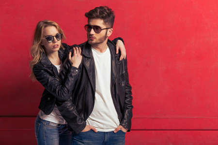 Cool beautiful young couple in leather jackets and sunglasses is looking at camera, standing against red backgroundの写真素材