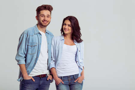 Beautiful young couple in jeans is smiling and looking at camera, standing, hands in pockets, on a gray backgroundの写真素材
