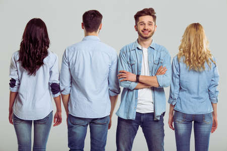 Young people in jeans on a gray background. Three are standing turned back, one man is looking at camera and smilingの写真素材