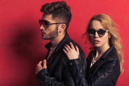 Portrait of beautiful young couple in leather jackets and sunglasses standing against red...