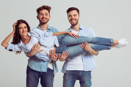 Beautiful young people in jeans are looking at camera and smiling, on a gray background. Two men are holding woman on handsの写真素材