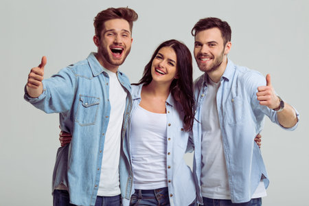 Beautiful young people in jeans are showing Ok sign, looking at camera and smiling, on a gray...