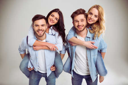 Two beautiful young couples in jeans are looking at camera and smiling, standing on a gray background. Girls pickabackの写真素材