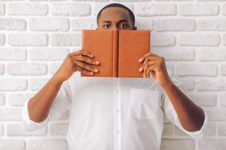 Handsome Afro American man in classic shirt is covering half face with a notebook, standing against...