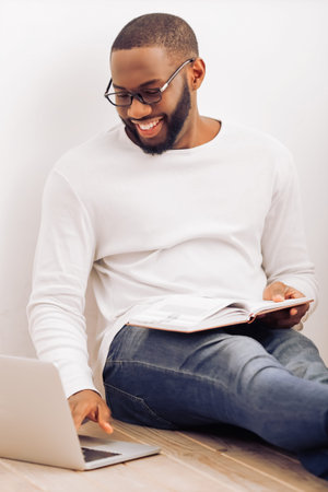 Handsome Afro American man in glasses is making notes, using a laptop and smiling while sitting on...