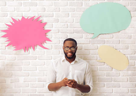 Handsome Afro American man in classic shirt and glasses is using a smart phone, standing with speech bubbles around him against brick wallの写真素材