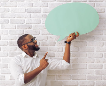 Handsome Afro American man in classic shirt and glasses is holding a speech bubble and smiling,...