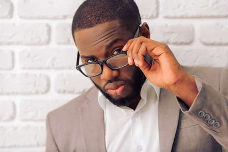 Handsome Afro American man in classic suit is looking at camera from under his glasses, standing against white brick wallの写真素材