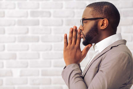 Side view of handsome Afro American man in classic suit and glasses keeping palms together like praying, standing against white brick wall