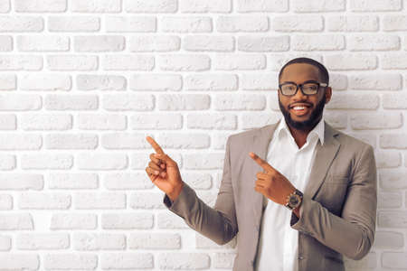 Handsome Afro American man in classic suit and glasses is smiling, looking at camera and pointing away, against white brick wallの写真素材