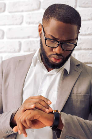 Handsome Afro American man in classic suit and glasses looking at his watch, against white brick wall, close upの写真素材