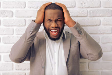 Stressed Afro American man in classic suit is keeping hands on head and screaming, against white brick wallの写真素材