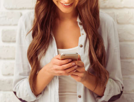 Beautiful young woman is using a smart phone and smiling, against white brick wall, croppedの写真素材