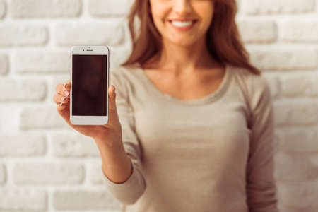 Beautiful young woman in casual wear is presenting a smartphone, looking at camera and smiling, standing against white brick wall, close upの写真素材