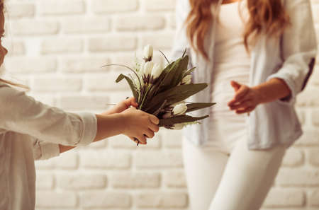 Cute little girl is giving flowers to her beautiful young mother, against white brick wall, close upの写真素材