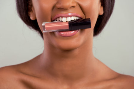 Portrait of beautiful Afro American girl holding a lipstick in her teeth, on a gray background,...