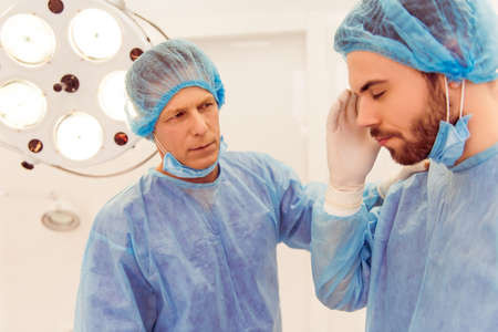 Handsome young doctor is having a headache, his colleague is calming him, standing in a modern operating roomの写真素材