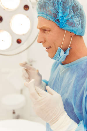 Side view of handsome middle aged doctor keeping fingers crossed before the operation, standing in a modern operating roomの写真素材