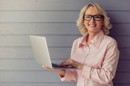Beautiful old woman in classic shirt and glasses is using a laptop, looking at camera and smiling,...