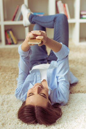 Beautiful young girl is using a smartphone and smiling, lying on her back on carpet at homeの写真素材