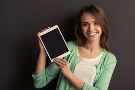 Portrait of beautiful young girl in casual clothes showing a tablet, looking at camera and smiling, against dark backgroundの写真素材