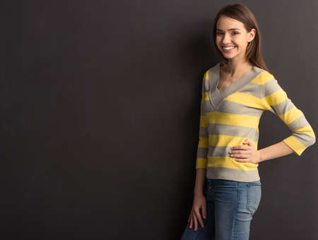 Beautiful young girl in casual clothes is looking at camera and smiling, standing against dark backgroundの写真素材