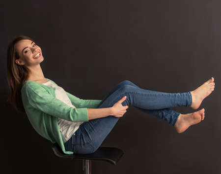 Side view of beautiful young girl looking at camera and smiling, sitting with raised legs on chair against dark backgroundの写真素材