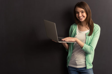 Beautiful young girl in casual clothes is using a laptop, looking at camera and smiling, standing against dark backgroundの写真素材
