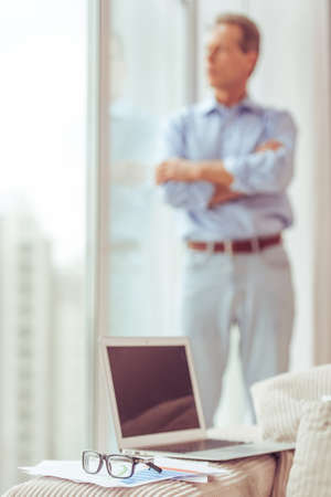 Handsome middle aged businessman in classic shirt is looking out the window, a laptop in focus in the foregroundの写真素材