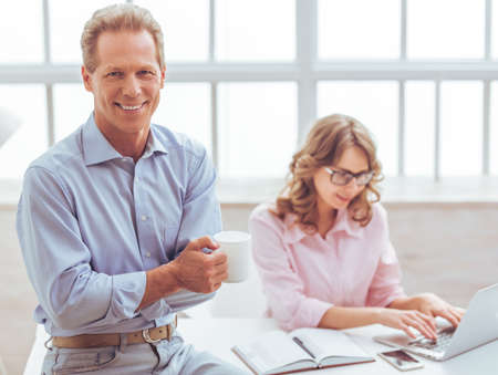 Handsome businessman is holding a cup and smiling, in the background business woman working using a laptopの写真素材