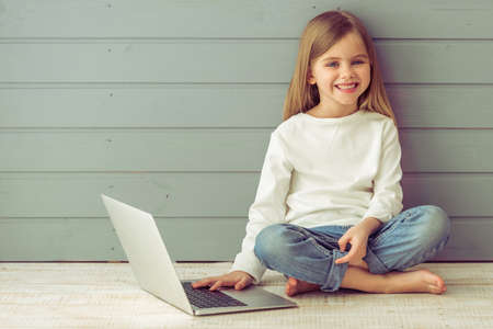 Pretty little girl in casual clothes is using a laptop, looking at camera and smiling, against gray backgroundの写真素材