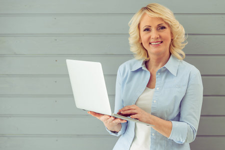 Beautiful mature woman in casual clothes is using a laptop, looking at camera and smiling, standing against gray backgroundの写真素材