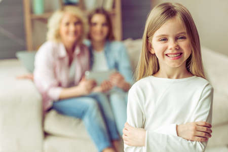 Little girl is looking at camera and smiling, in the background her mom and granny are using a tablet while sitting on sofa at homeの写真素材