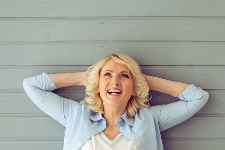 Portrait of beautiful mature woman looking up and smiling, standing with hands behind head against...