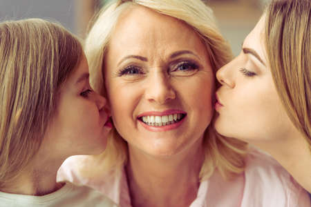 Beautiful granny is looking at camera and smiling while her daughter and granddaughter are kissing her