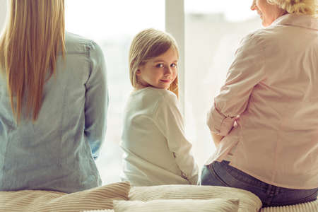 Back view of three generations of beautiful women sitting on sofa against window. Little girl looking at camera and smilingの写真素材