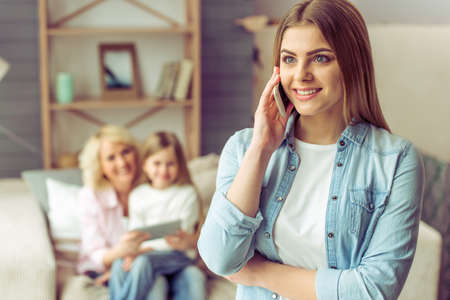 Young woman is talking on the mobile phone, in the background her mother and daughter are using a tablet, sitting on sofa at homeの写真素材